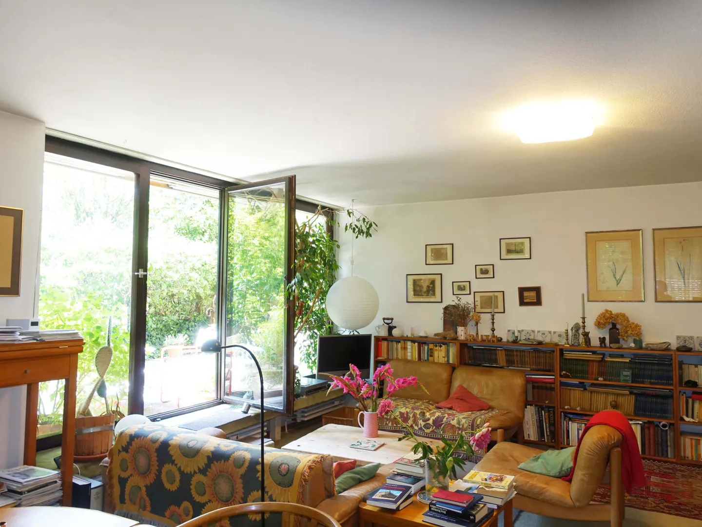 Bright living room with large windows overlooking a green garden. Bookshelves line the wall, and a brown leather sofa sits in the center.