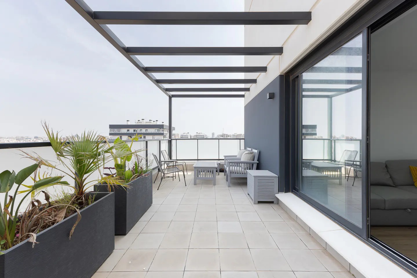 Modern balcony with gray furniture, planters, and a pergola. Sliding glass doors lead indoors. City view in the background.