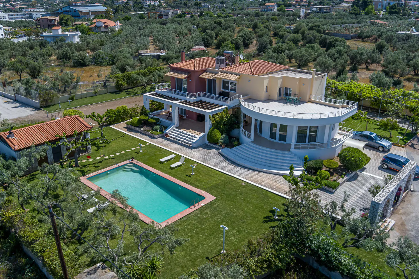 Aerial view of a two-story house with a pool, green lawn, and trees in the background. The house has a red tile roof and a white exterior.