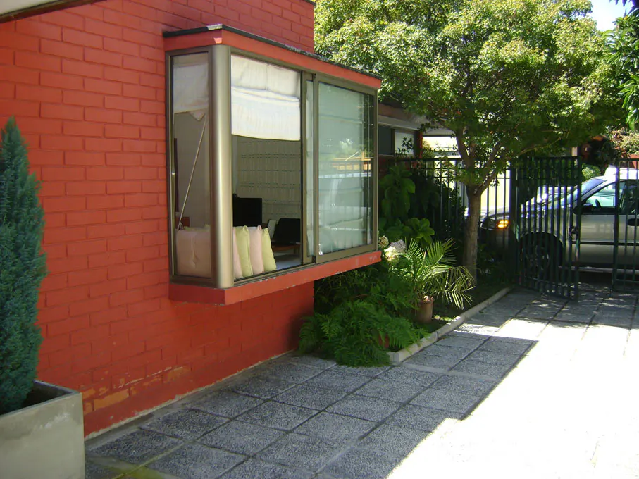 Exterior of a red brick house with a bay window, gray stone walkway, and a car parked behind a black metal gate.