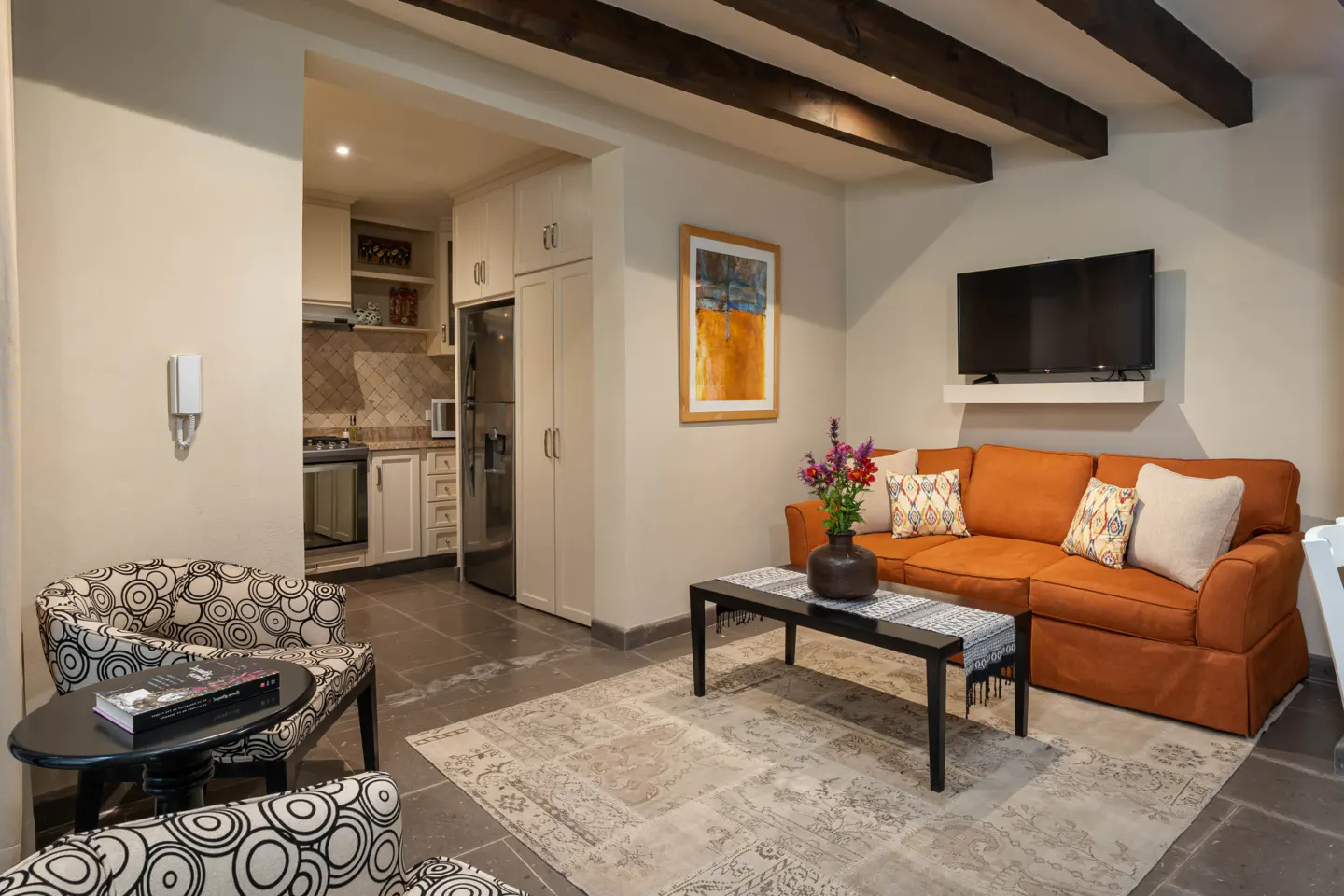 Living room with orange sofa, black coffee table, patterned chairs, and kitchen view. Dark wood beams on ceiling.