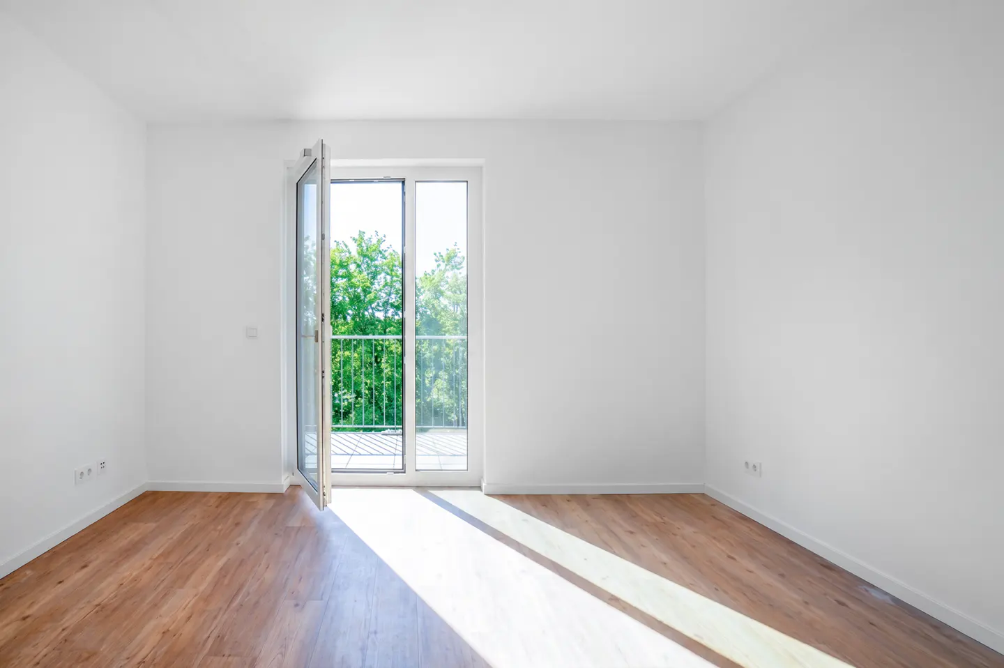 Bright, empty room with wood floors and white walls. Open glass doors lead to a balcony with green trees visible.