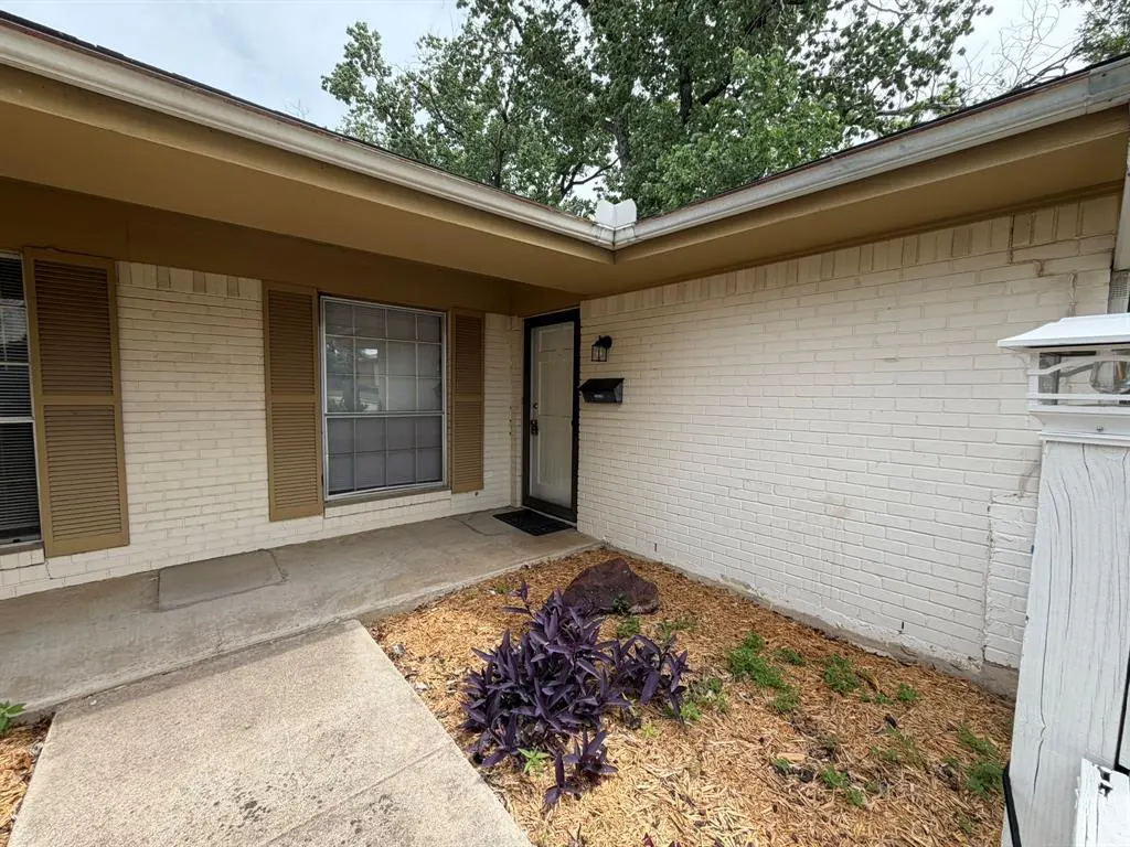 Exterior of a one-story white brick house with brown shutters and a black front door. A small garden bed is in front.