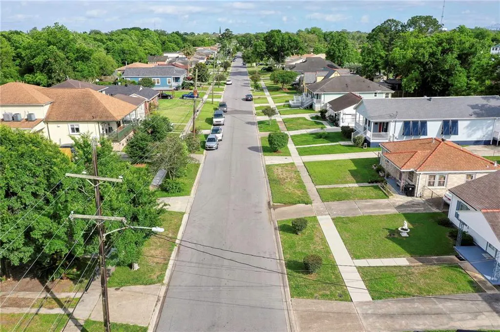 Aerial view of a quiet residential street lined with houses, green lawns, and parked cars under a partly cloudy sky.