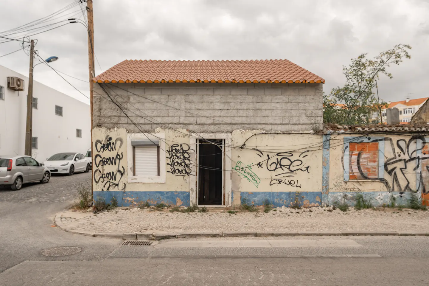 A small, graffiti-covered building with a red tile roof sits on a street corner under a cloudy sky.