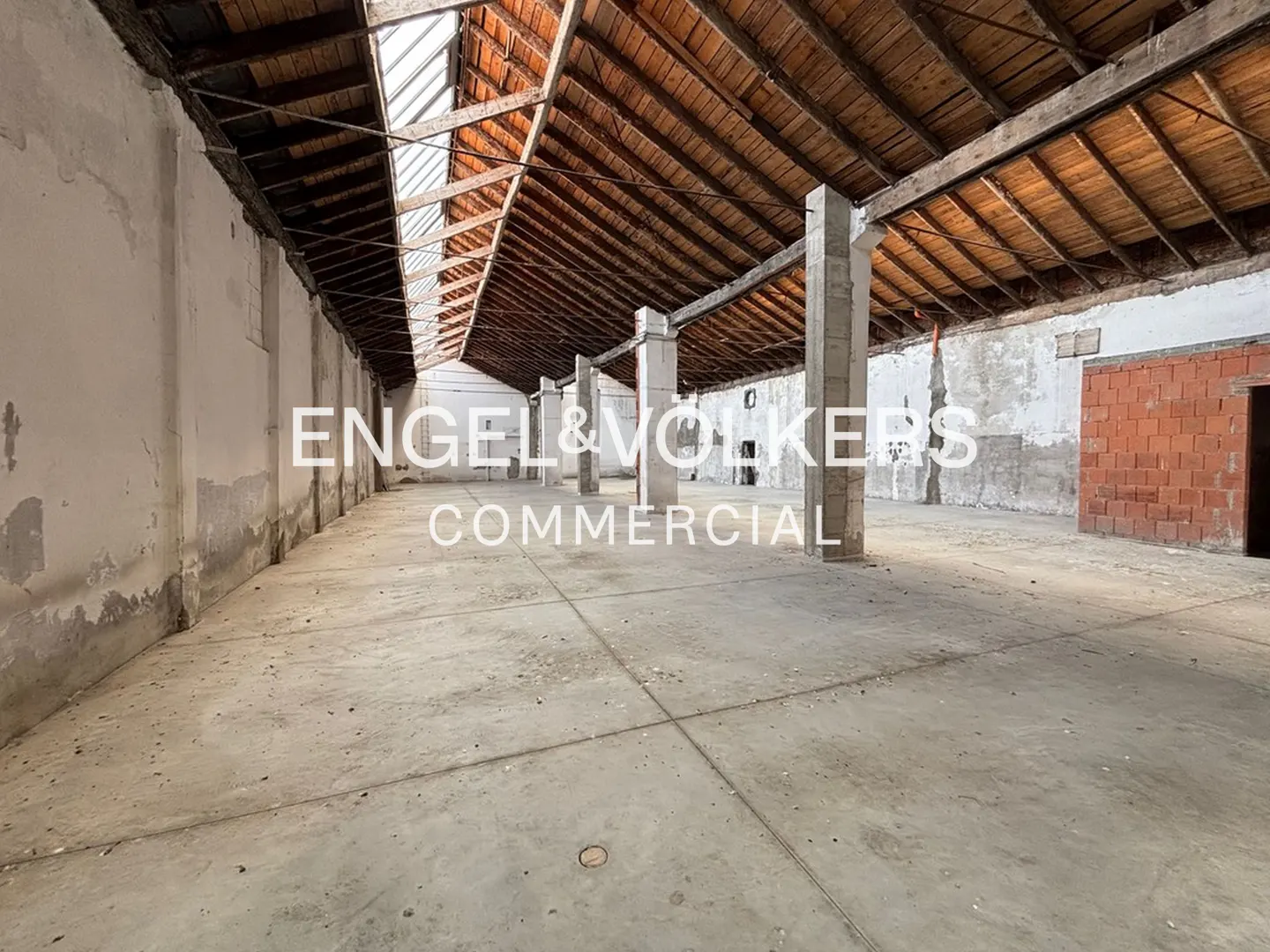 An empty commercial space with concrete floors, white walls, and a wooden ceiling with skylights. Engel & Volkers logo is visible.