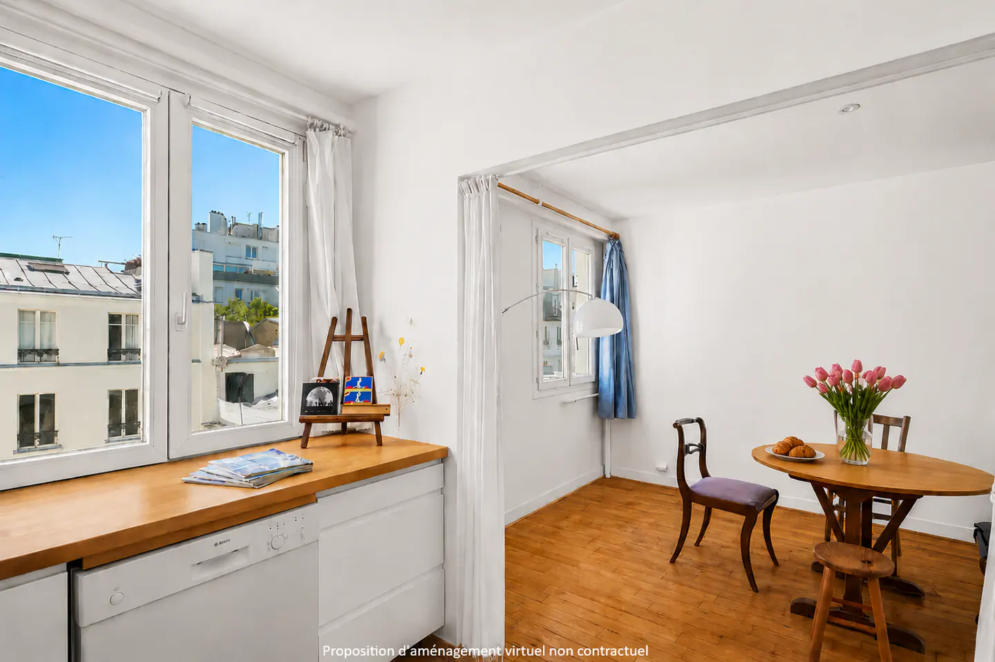 Bright apartment interior with white walls, wood floors, and a view of Paris. A table with tulips and pastries sits in the dining area.