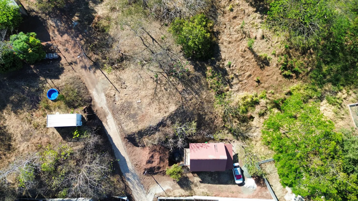 Aerial view of a property with a red-roofed house, a white car, and a dirt road surrounded by trees and vegetation.