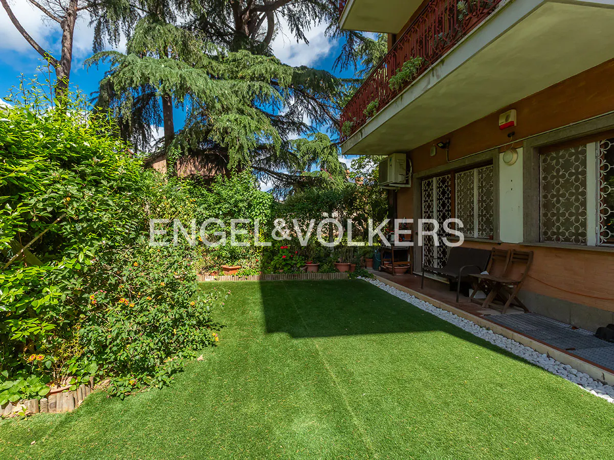 A lush green lawn with trees and bushes leads to a two-story house with a red balcony railing.