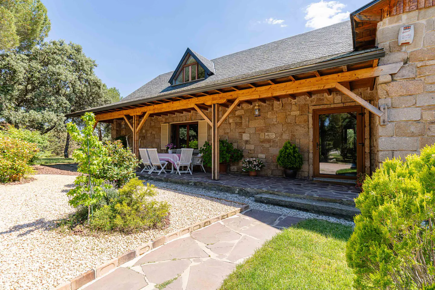 Stone house exterior with a covered porch, table, and chairs. A stone path leads to the house with green grass and trees.