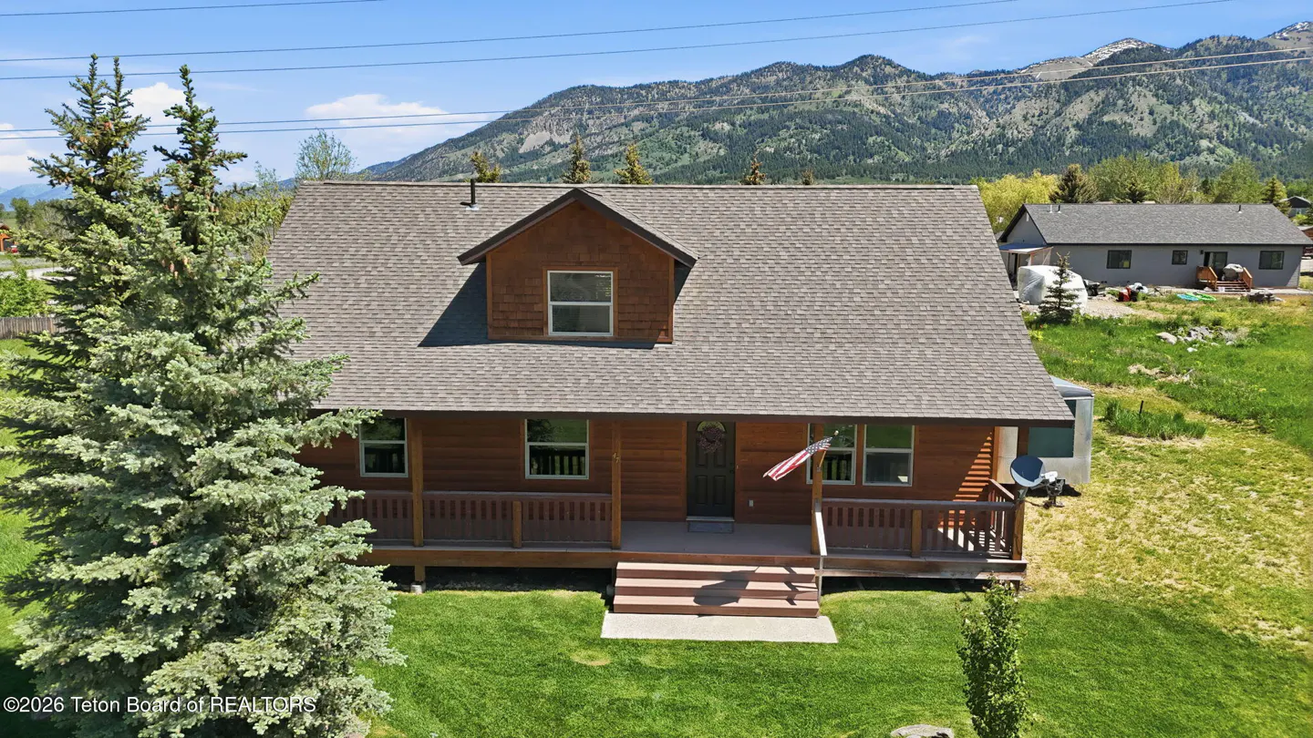 A two-story wooden cabin with a front porch and American flag, set against a backdrop of mountains and green lawn.