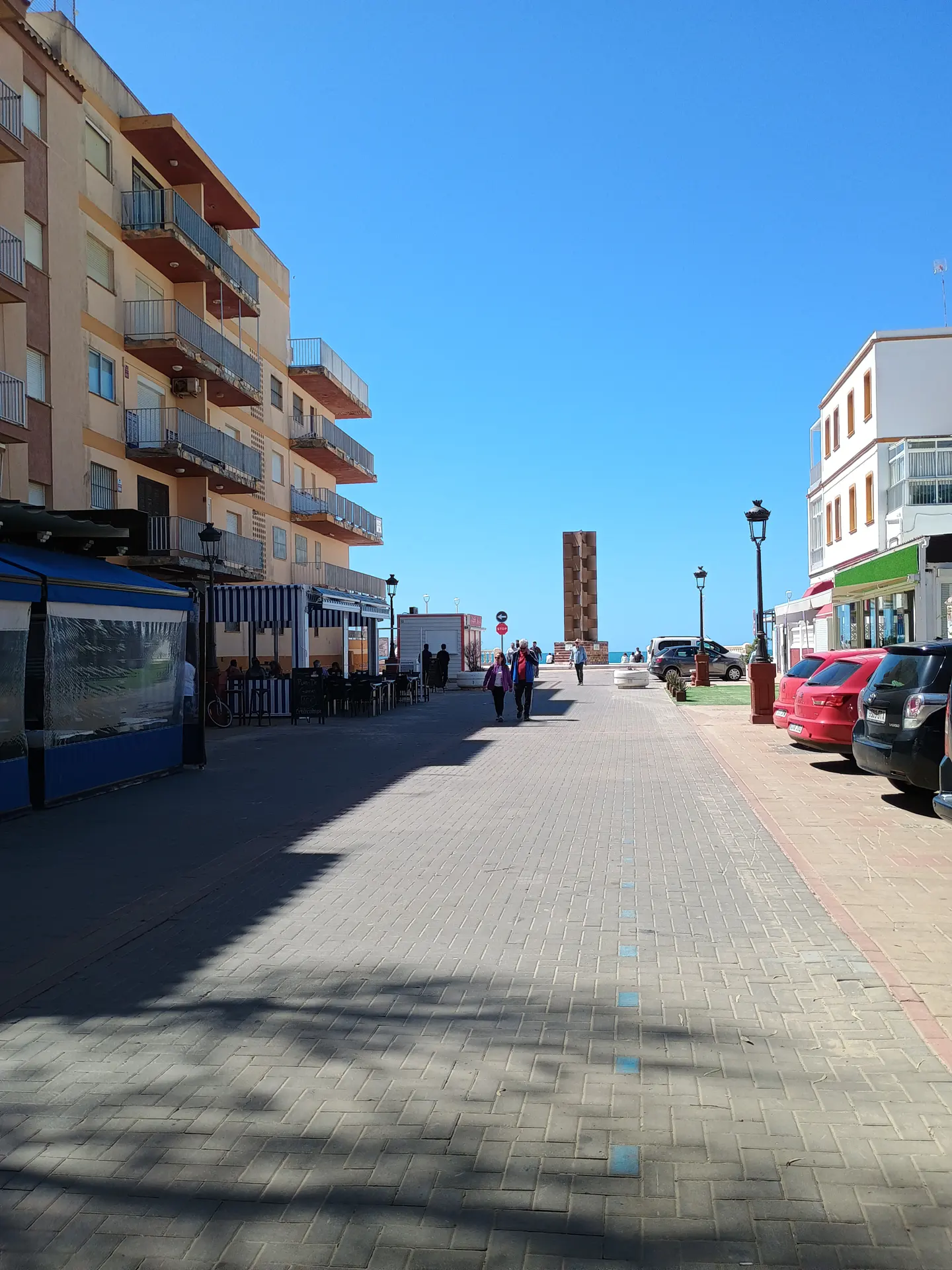 Coastal scene with a brick walkway, buildings, and parked cars under a clear blue sky. A monument stands in the distance.