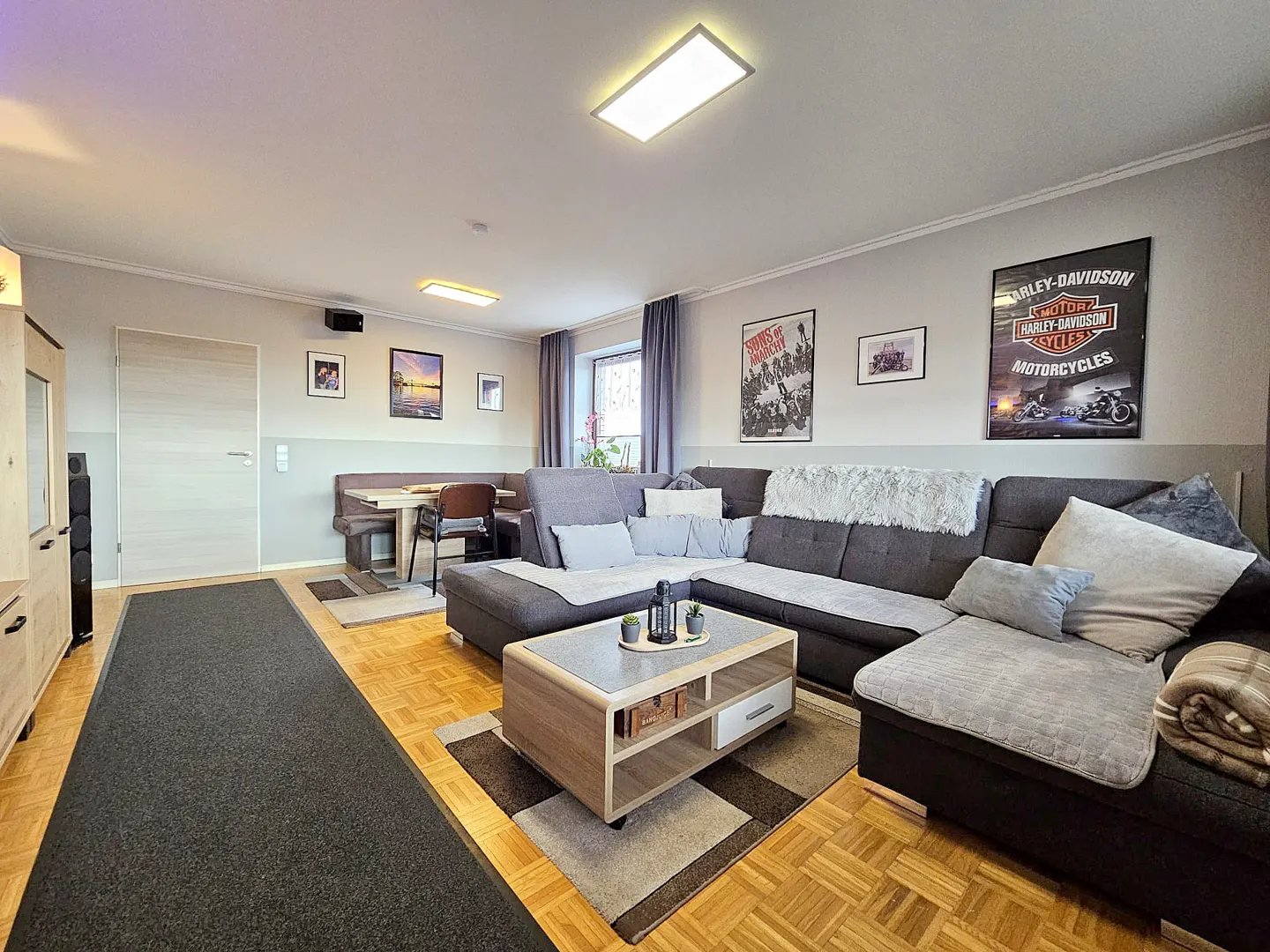 Living room with a gray sectional sofa, a wooden coffee table, and a dining area with a bench and table. Posters decorate the wall.