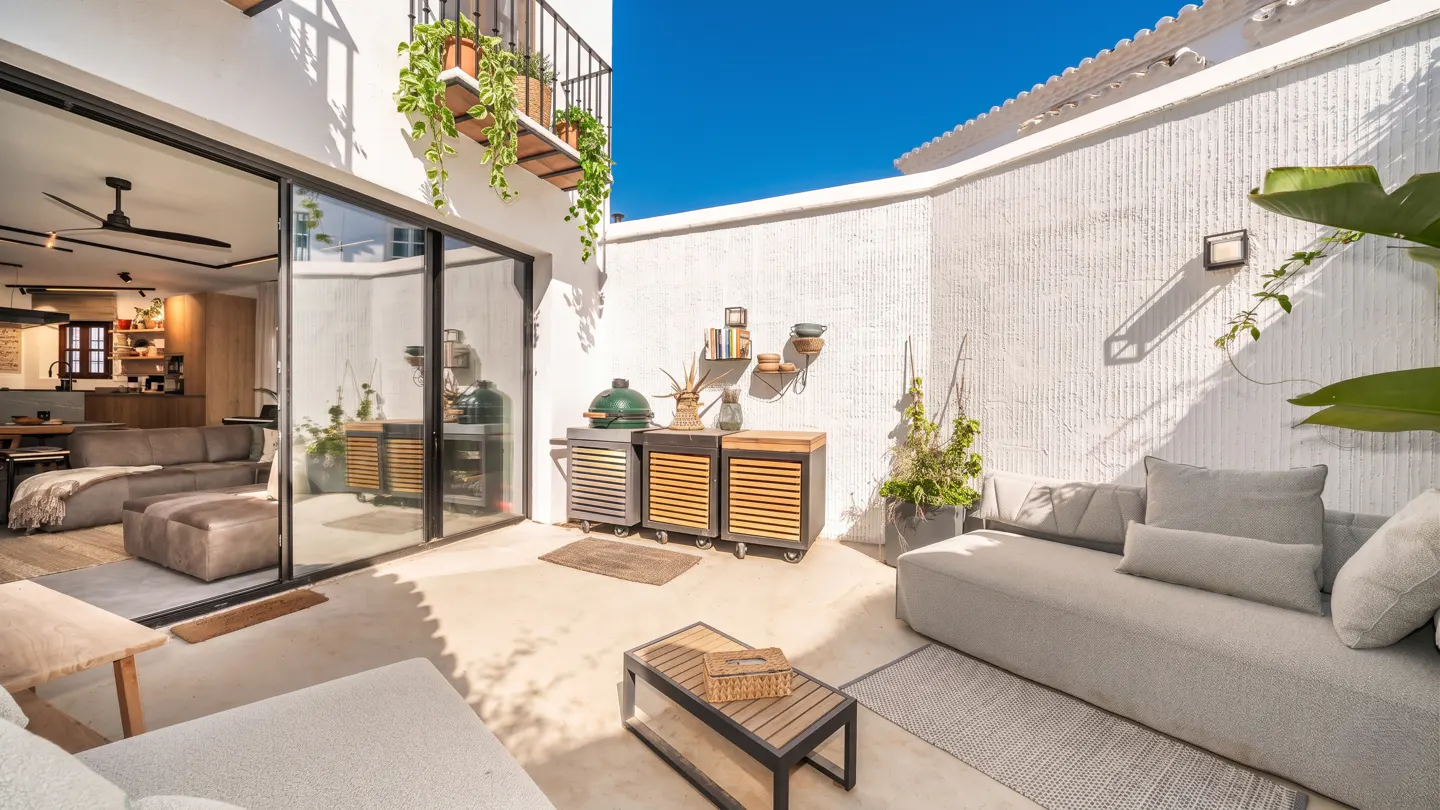 Outdoor patio with a gray sofa, wood table, and grill. Sliding glass doors lead to the living room. White walls and blue sky.