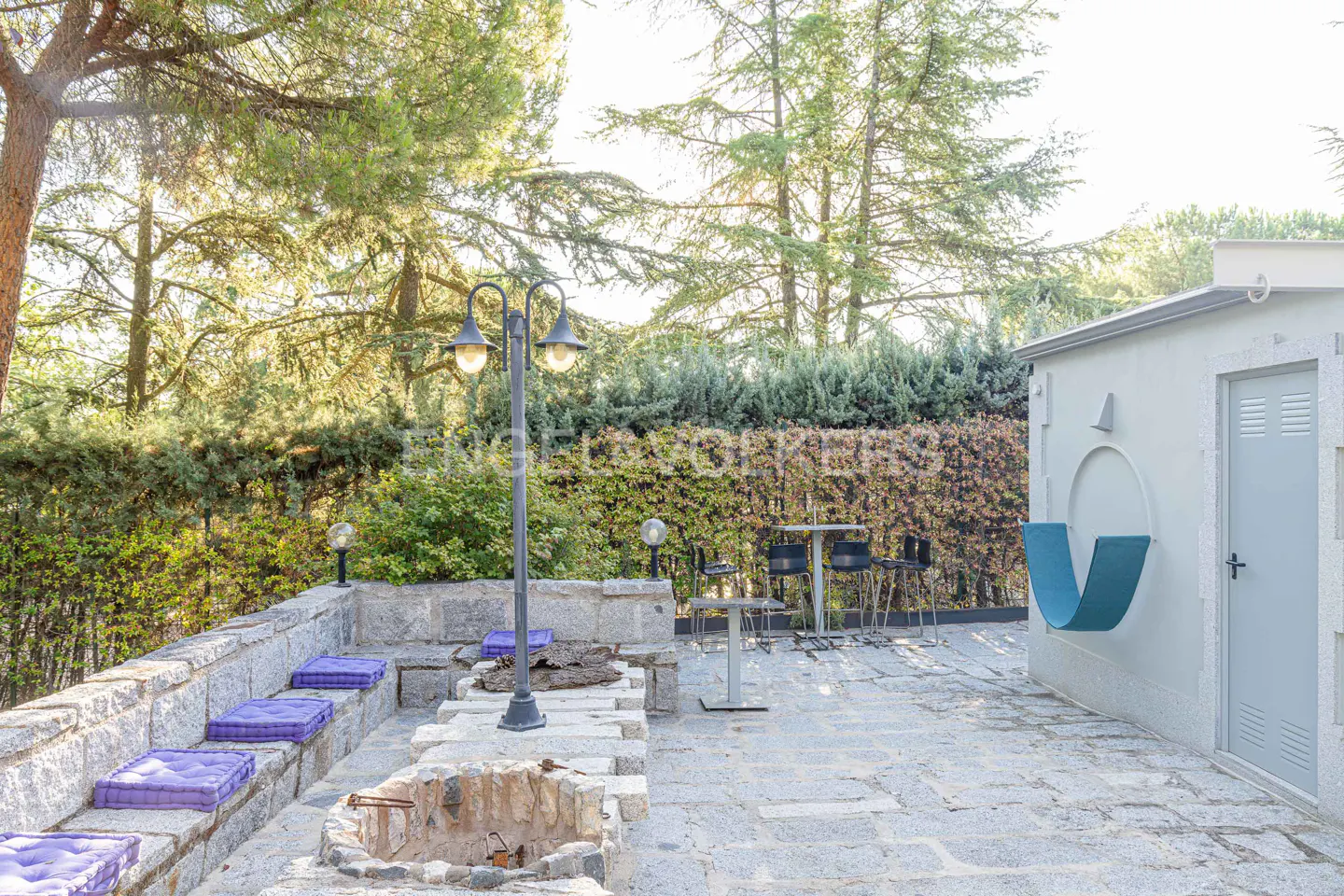 Outdoor patio with stone seating, purple cushions, fire pit, table with chairs, and a modern blue chair on a white building.