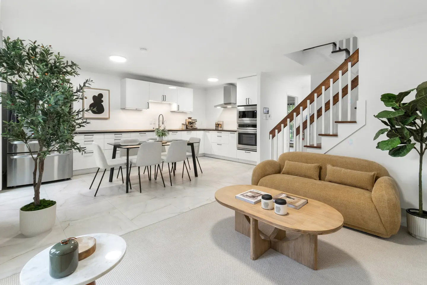 Open-plan kitchen and living area with white cabinets, a dining table, a tan sofa, and indoor plants.