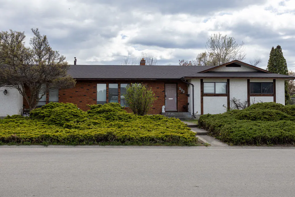A single-story house with a brick and stucco exterior, green bushes, and a dark roof.