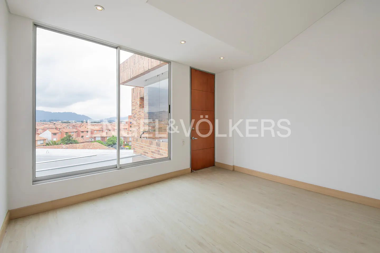 Bright, empty room with light wood floors, white walls, and a large window overlooking a cityscape. A wooden door is on the right.