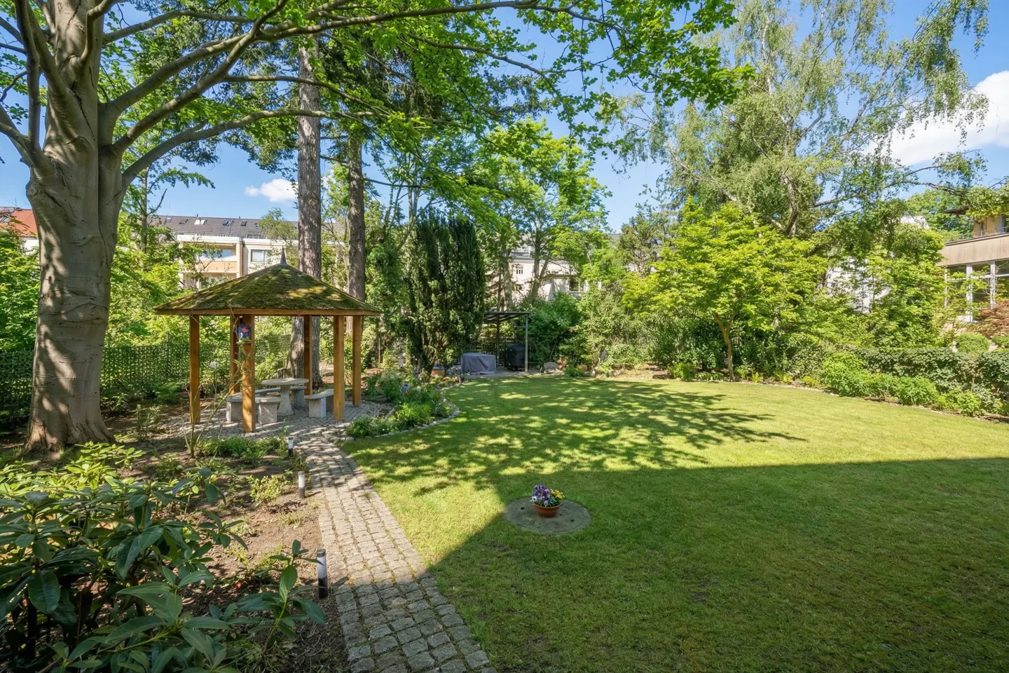 Lush green backyard with a stone path leading to a gazebo with a table and benches. Trees provide shade.