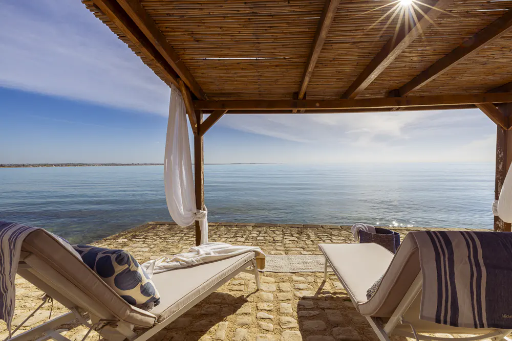 Ocean view from a cabana with two lounge chairs, blue pillows, and a bamboo roof.
