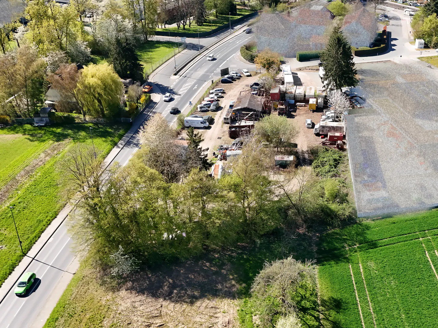 Aerial view of a property with a road, trees, and a field. Cars are driving on the road. There are buildings and parked vehicles on the property.