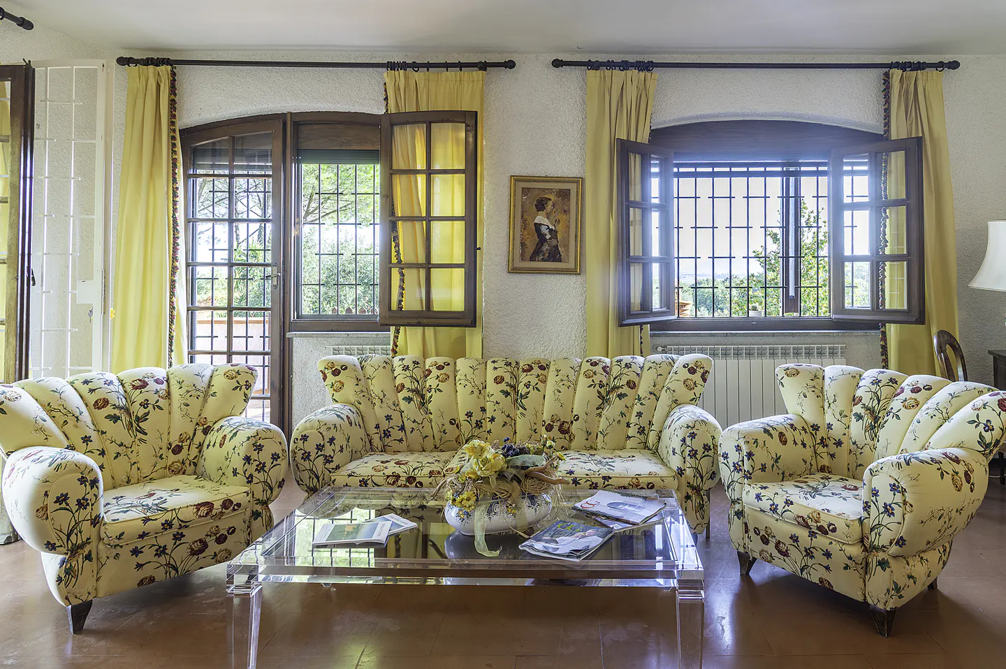 Living room with floral sofa and armchairs, clear coffee table, and open windows with yellow curtains.