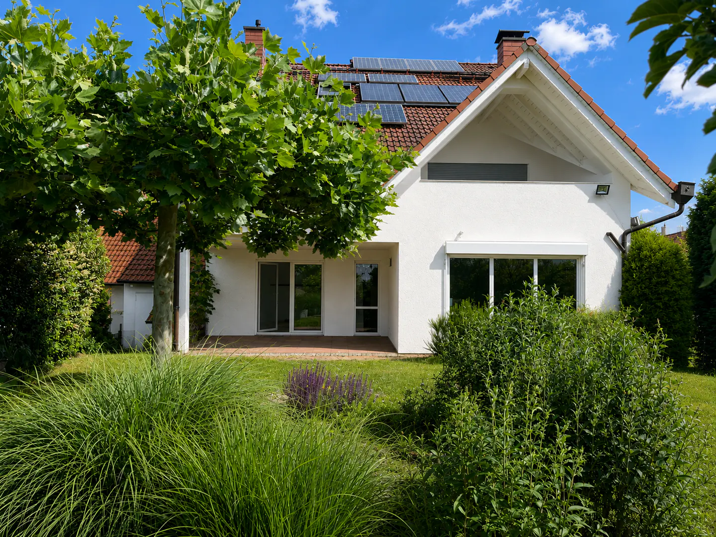 White house with solar panels on a tiled roof, featuring a patio, large windows, and a lush green garden under a blue sky.