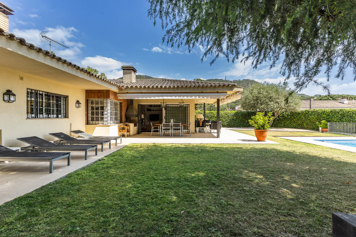 Backyard view of a tan house with a tiled roof, a covered patio with a table and chairs, a green lawn, and lounge chairs.