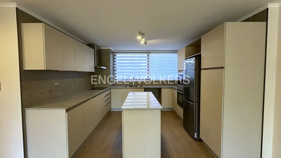 Modern kitchen with beige cabinets, granite countertops, a central island, stainless steel fridge, and window with blinds.