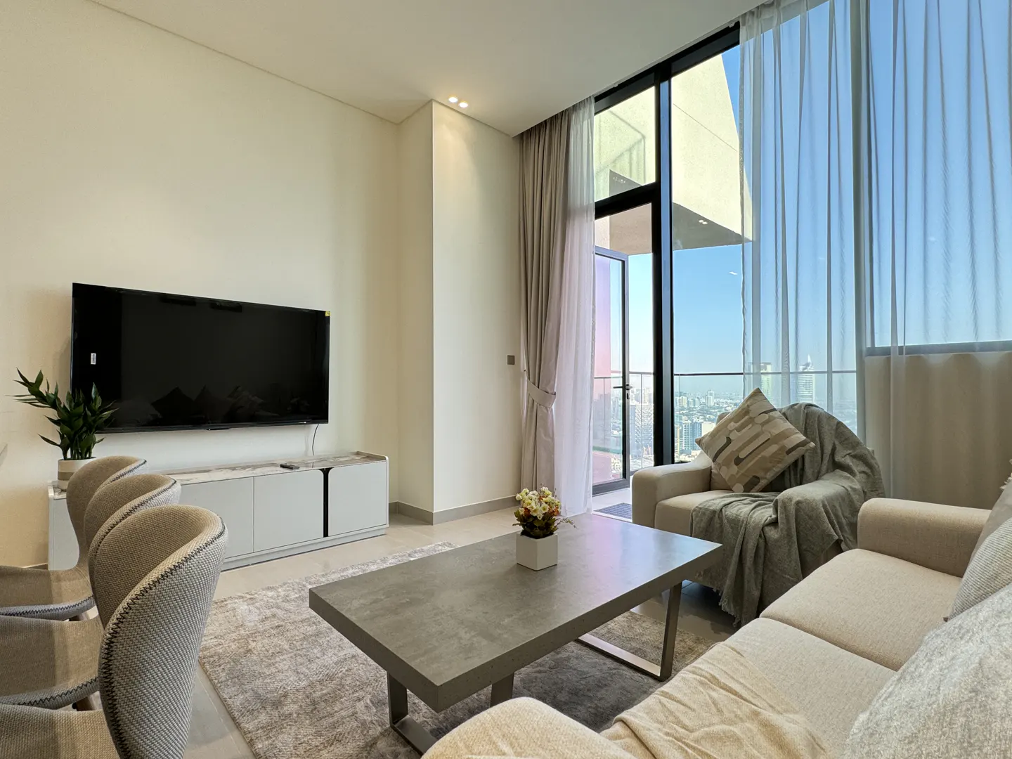 Living room with a TV, gray sofa, and a large window with city views. A gray coffee table sits on a gray rug.