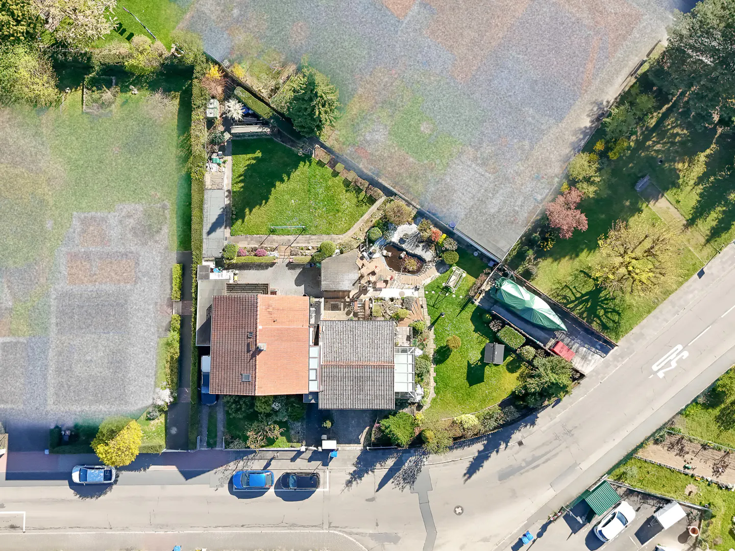 Aerial view of two houses with red and gray roofs, green lawns, a pond, and cars parked on the street.