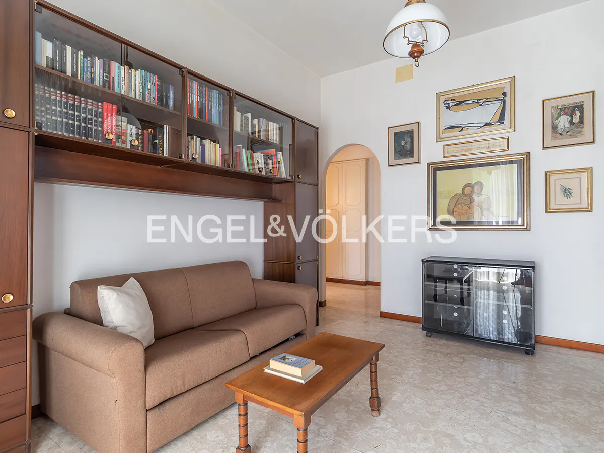 Living room with a brown sofa, wooden coffee table, and a large bookshelf filled with books. Artwork hangs on the white walls.