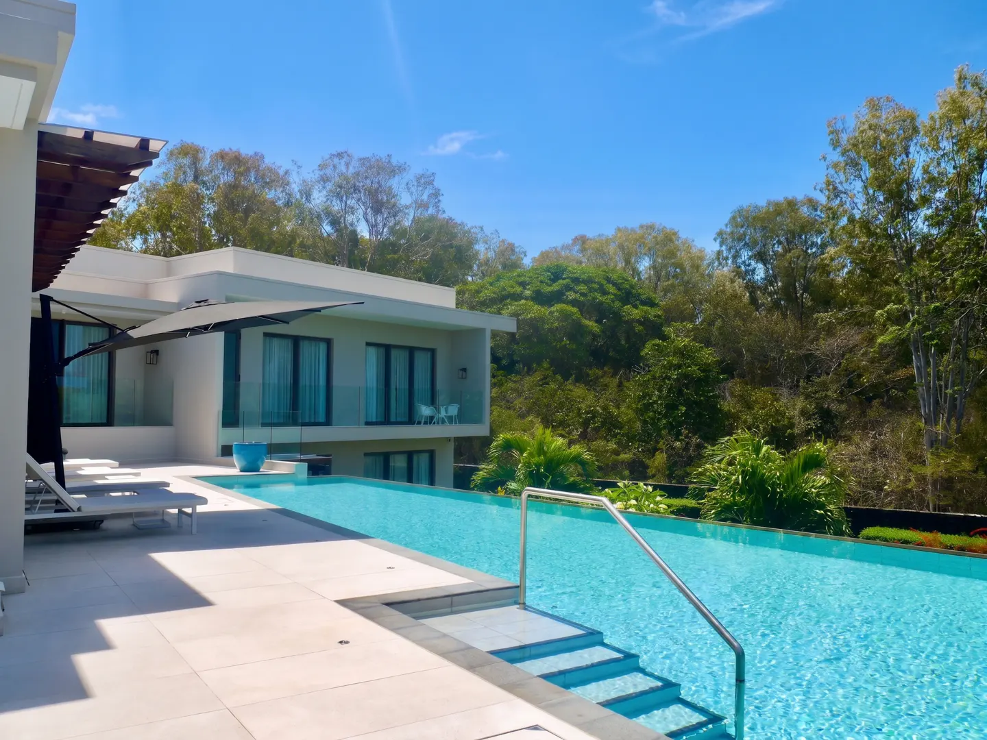 Luxury home with an infinity pool. The pool has steps and a handrail. Lounge chairs are on the patio. Trees are in the background.