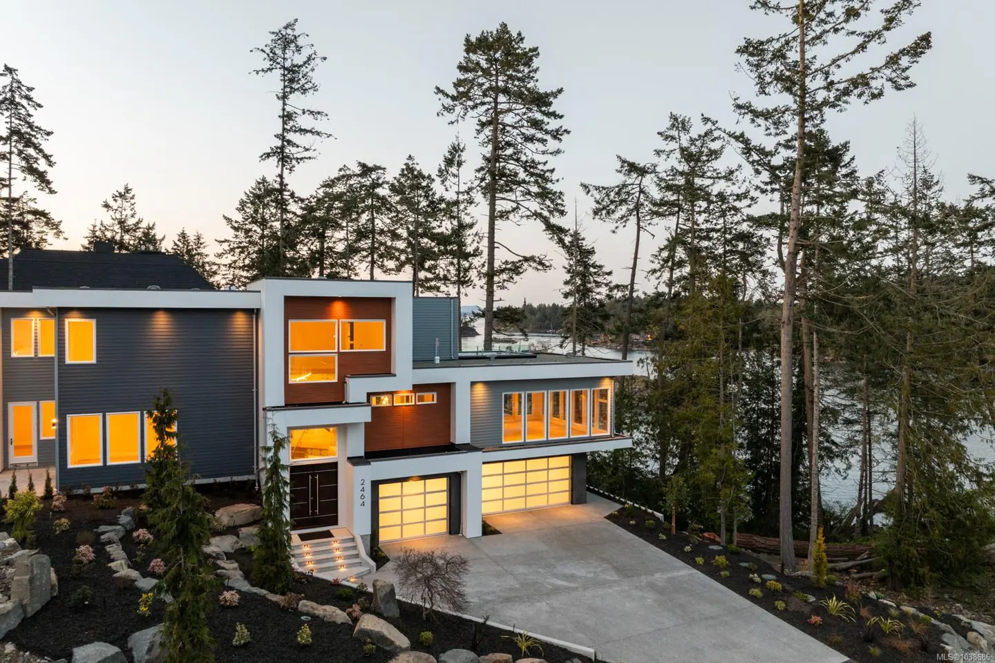 Modern two-story home with gray siding, brown accents, and a two-car garage. Large windows glow with warm light. Trees and water in background.