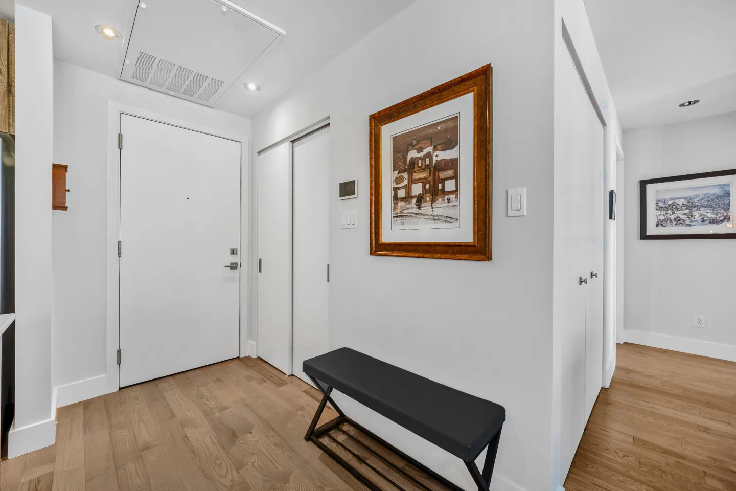 Entryway with a white door, wood floors, and a black bench under a framed picture on a white wall.