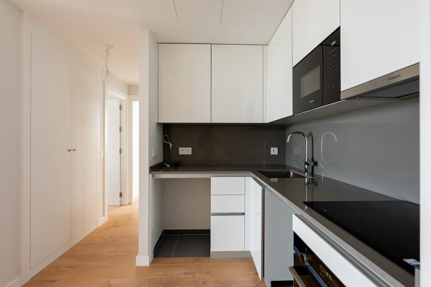 A modern kitchen with white cabinets, a black countertop, and stainless steel appliances. A hallway with white doors and wood floors is visible in the background.