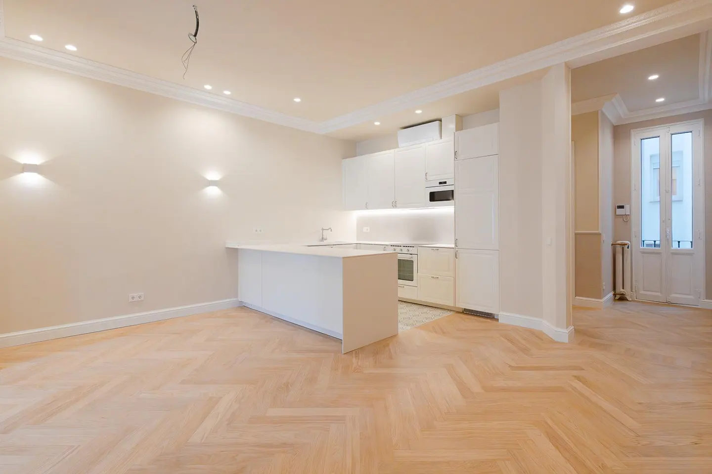 Bright, modern apartment with herringbone wood floors. White kitchen with island, cabinets, and appliances. Double doors lead to another room.