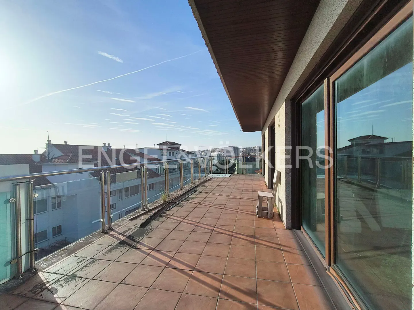 Wide balcony with glass railings and brown tile floor. A small white stool sits near large sliding glass doors. Cityscape visible in the background.