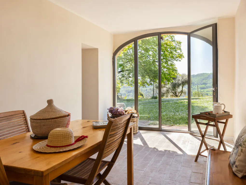Sunlit dining room with a wooden table, chairs, and a straw hat. Large arched windows offer a view of a green landscape.