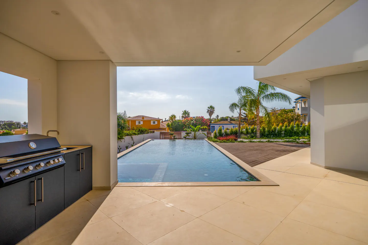 Outdoor kitchen with a black grill, sink, and cabinets. A pool is visible through the opening, with houses and palm trees in the background.