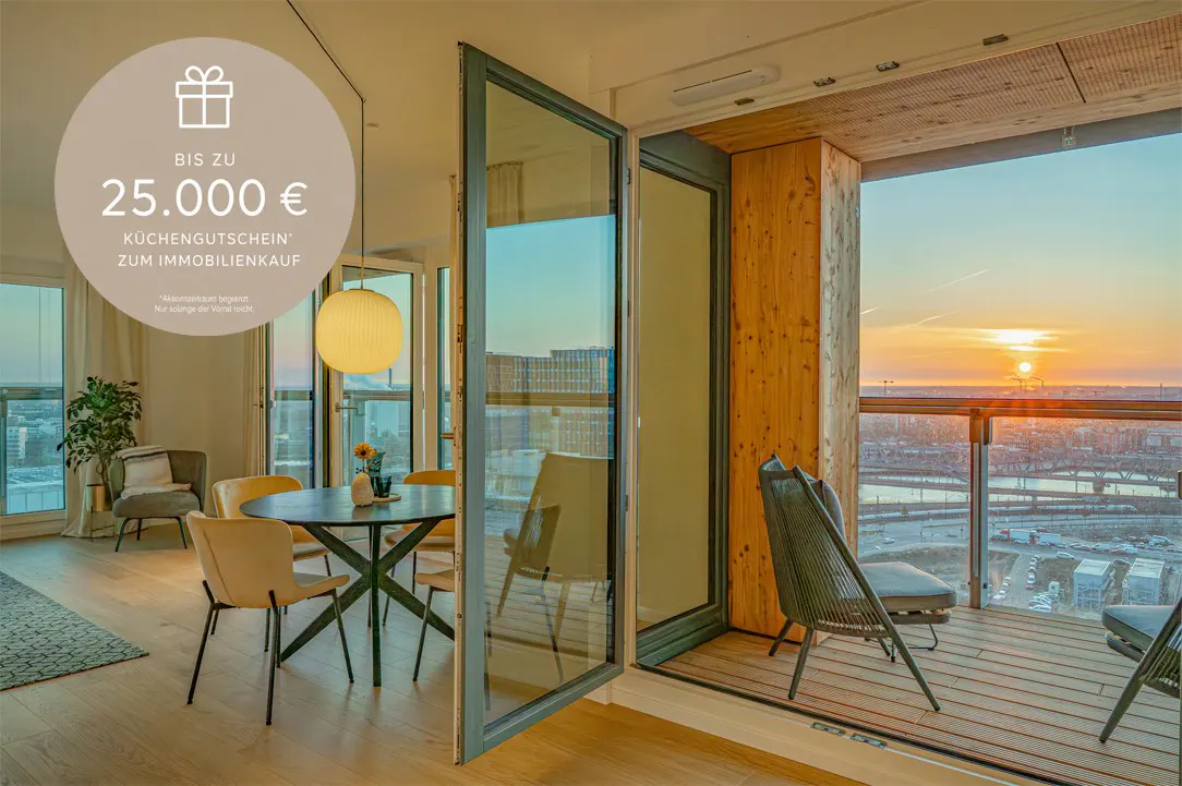 Bright apartment interior with open doors to a balcony overlooking a sunset cityscape. Dining table and chairs are visible.