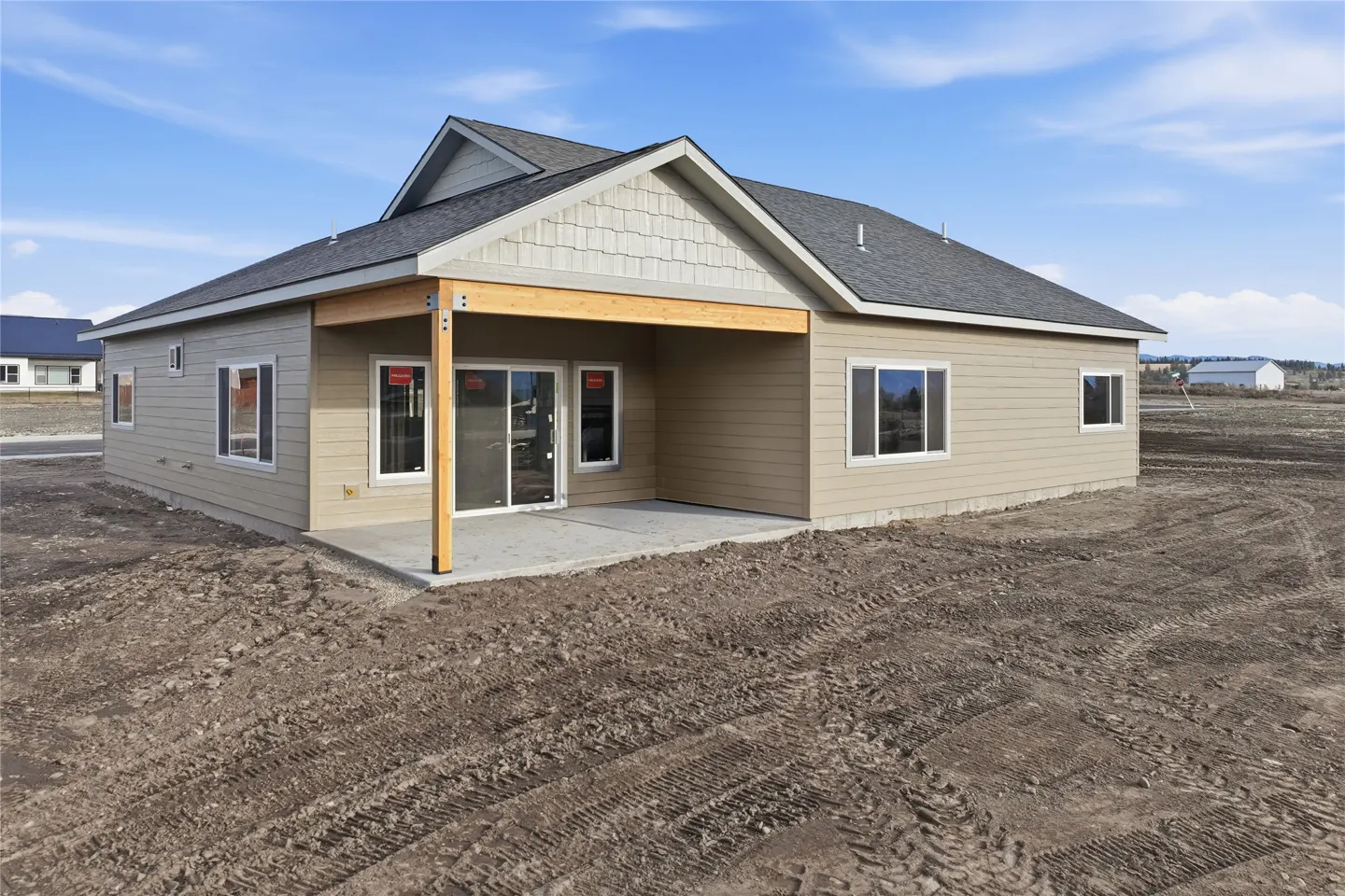 Rear exterior of a beige single-story home with a gray roof and a concrete patio. The yard is dirt with tire tracks. Blue sky.