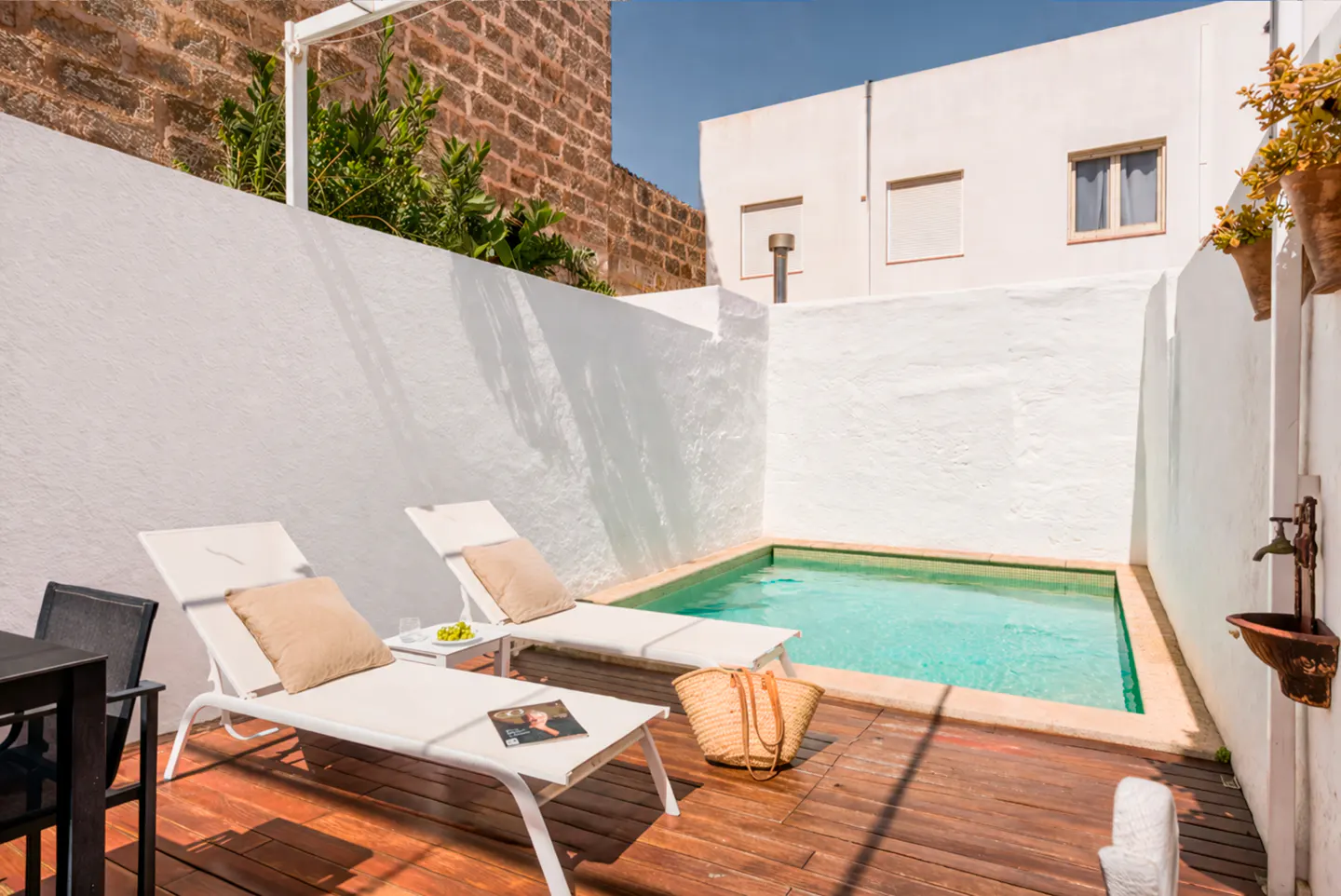 Outdoor patio with a small pool, two white lounge chairs with tan pillows, and a straw bag on a wooden deck. White walls surround the area.