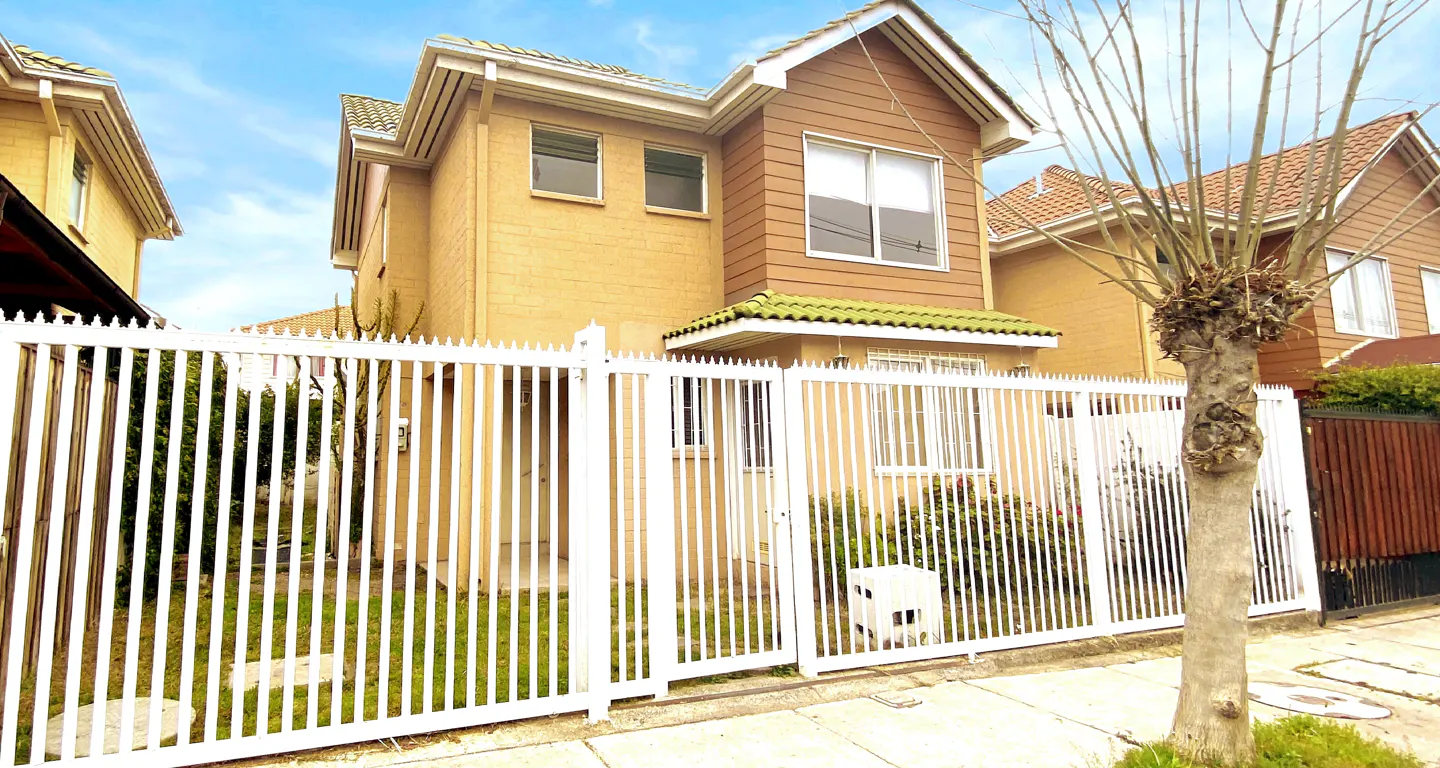 Two-story tan house with white trim and a green tile roof, surrounded by a white picket fence. A tree stands to the right.