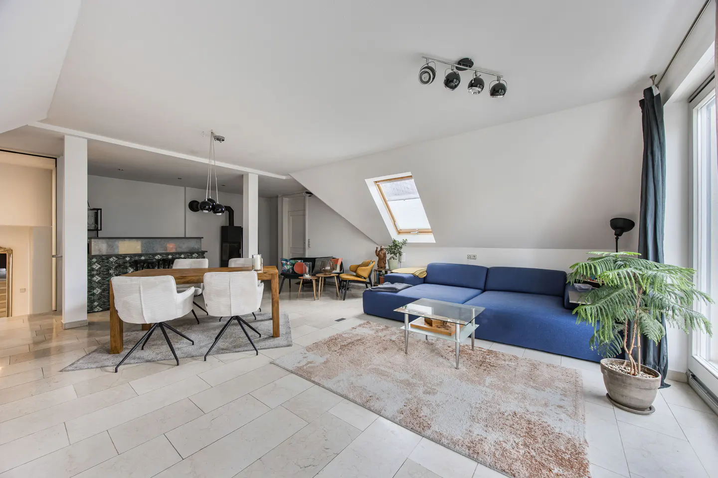 Bright, open-concept living space with a blue sofa, wood table with white chairs, and a large window. A potted plant sits near the window.