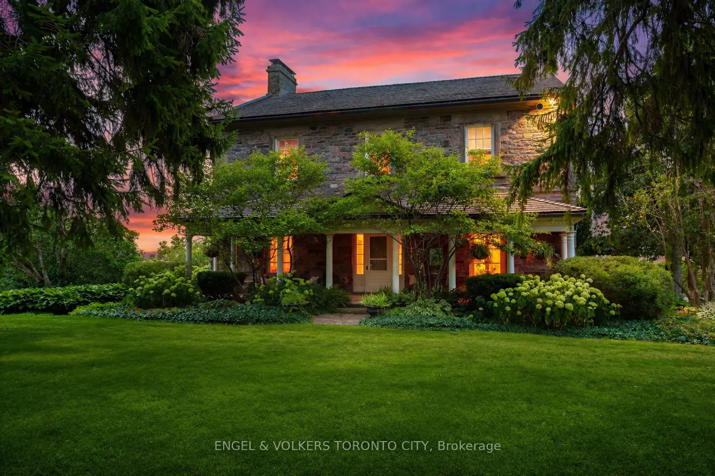 Exterior view of a two-story stone house with a porch, green lawn, and trees at sunset.