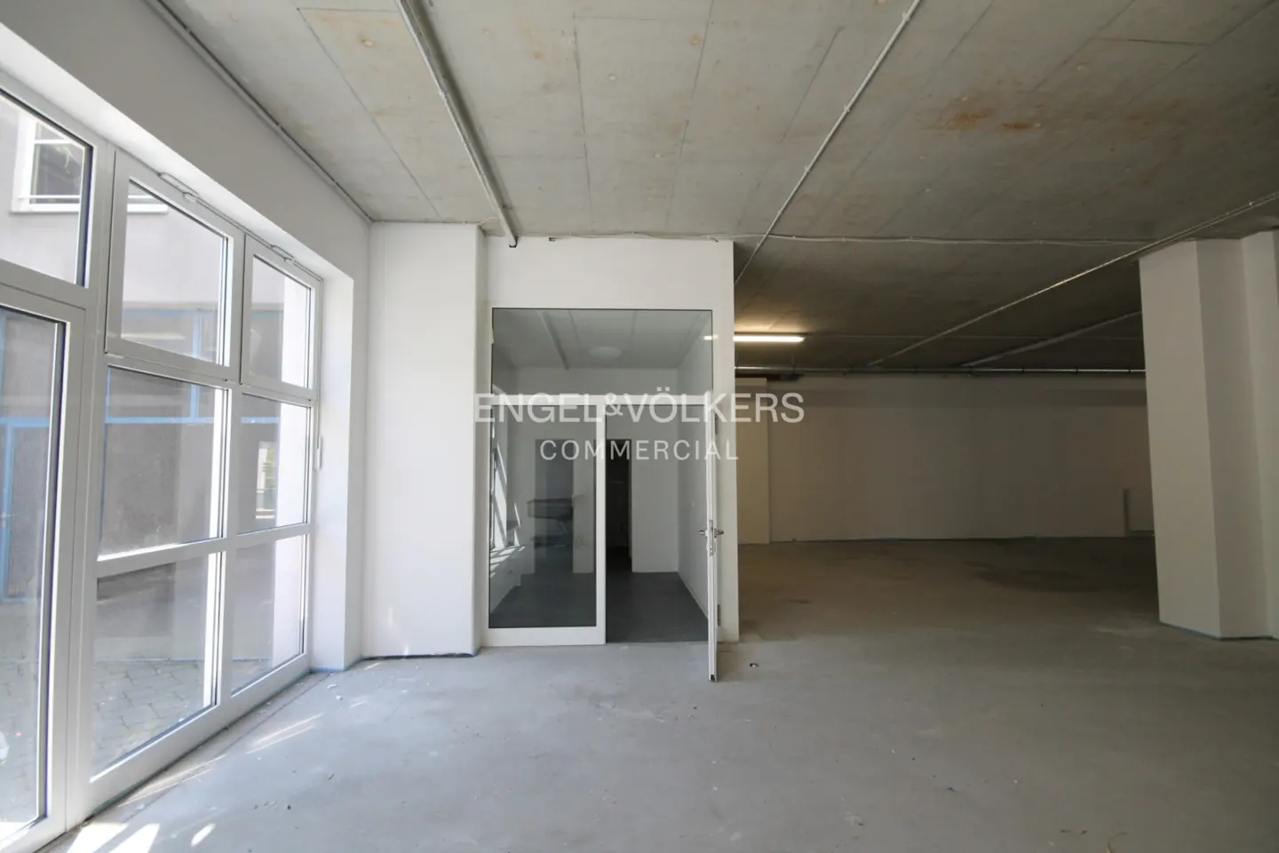 An empty, unfinished commercial space with concrete floors, white walls, a raw ceiling, and a glass-paneled office door.