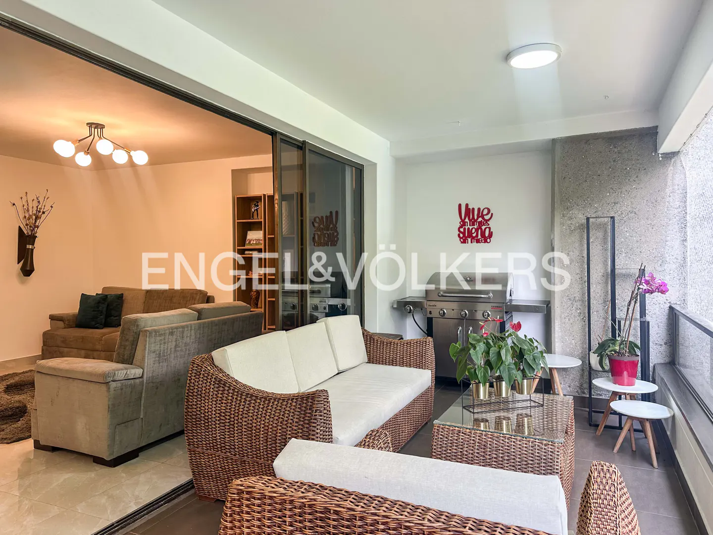 A bright, open-concept living space with a balcony featuring wicker furniture, a grill, and potted plants.