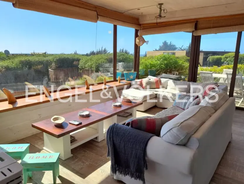 Sunroom with a white sofa, wood floors, and a long table. Large windows offer a view of green foliage and a blue sky.