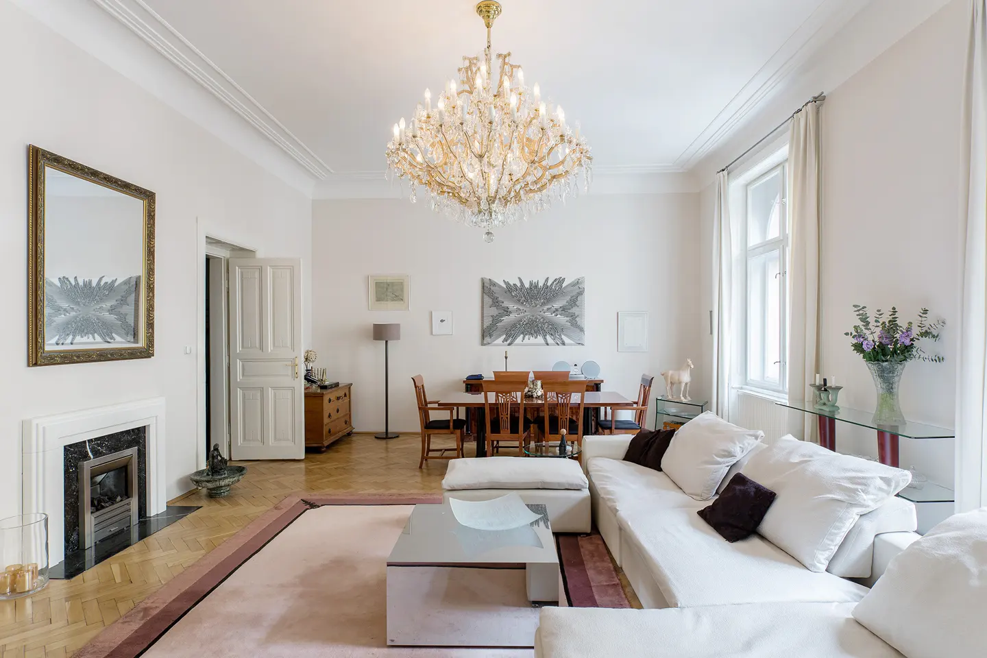Bright living room with white walls, a large crystal chandelier, a white sofa, and a dining table with chairs.